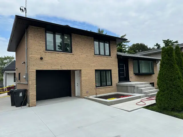 Front of a two-storey tan brick house with a black garage door, before painting
